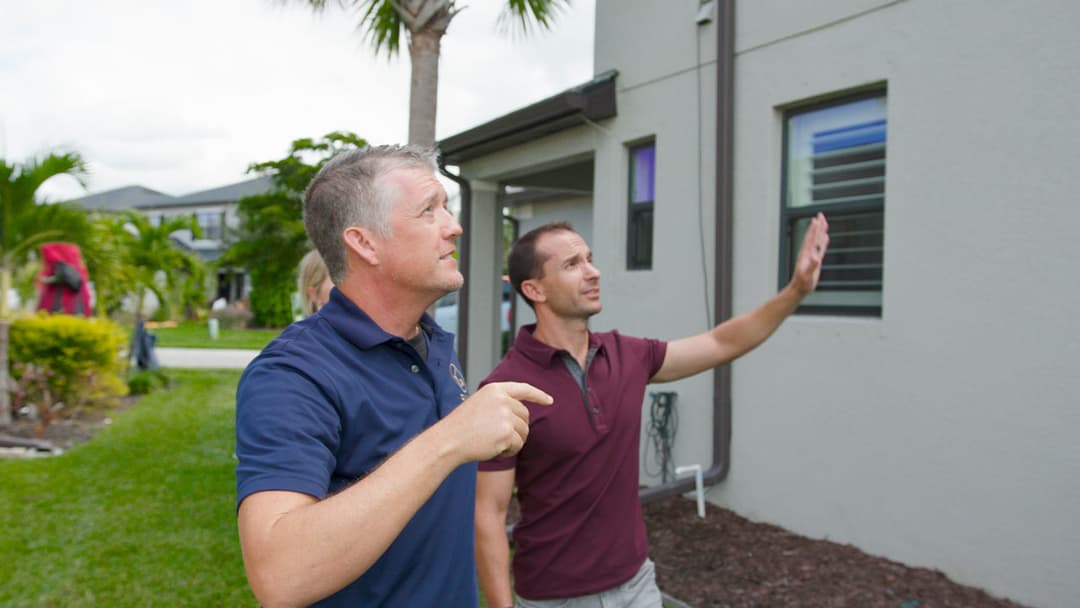 Two men inspecting a house exterior, discussing maintenance under palm trees.