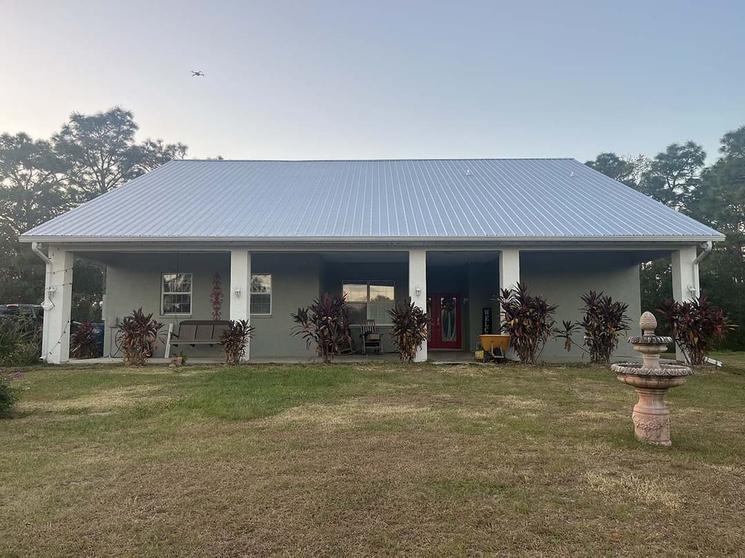 Single-story house with a metal roof, porch, and landscaped garden, surrounded by greenery.
