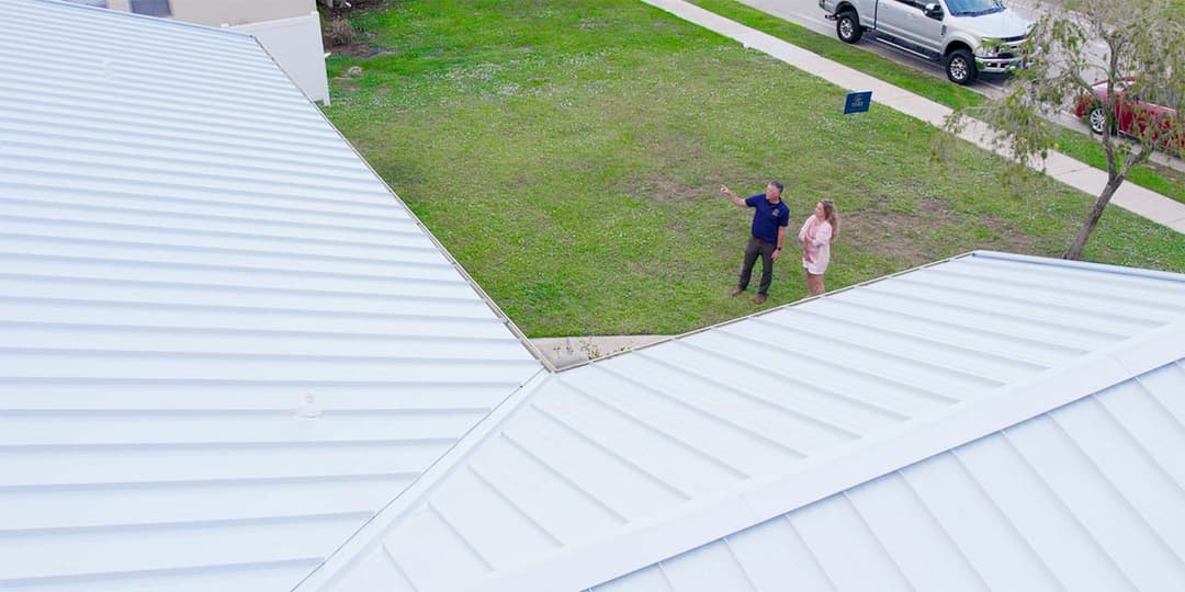 Two people discussing home exterior on a grassy lawn with a metal roof in view.