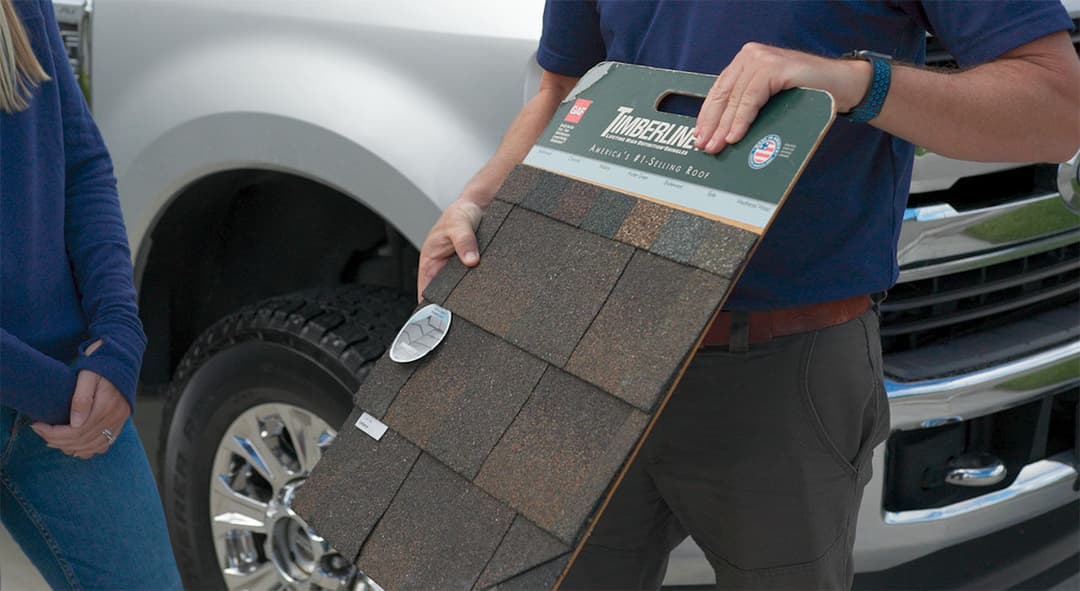 Person holding a Timberline roofing sample next to a vehicle, showcasing color options.