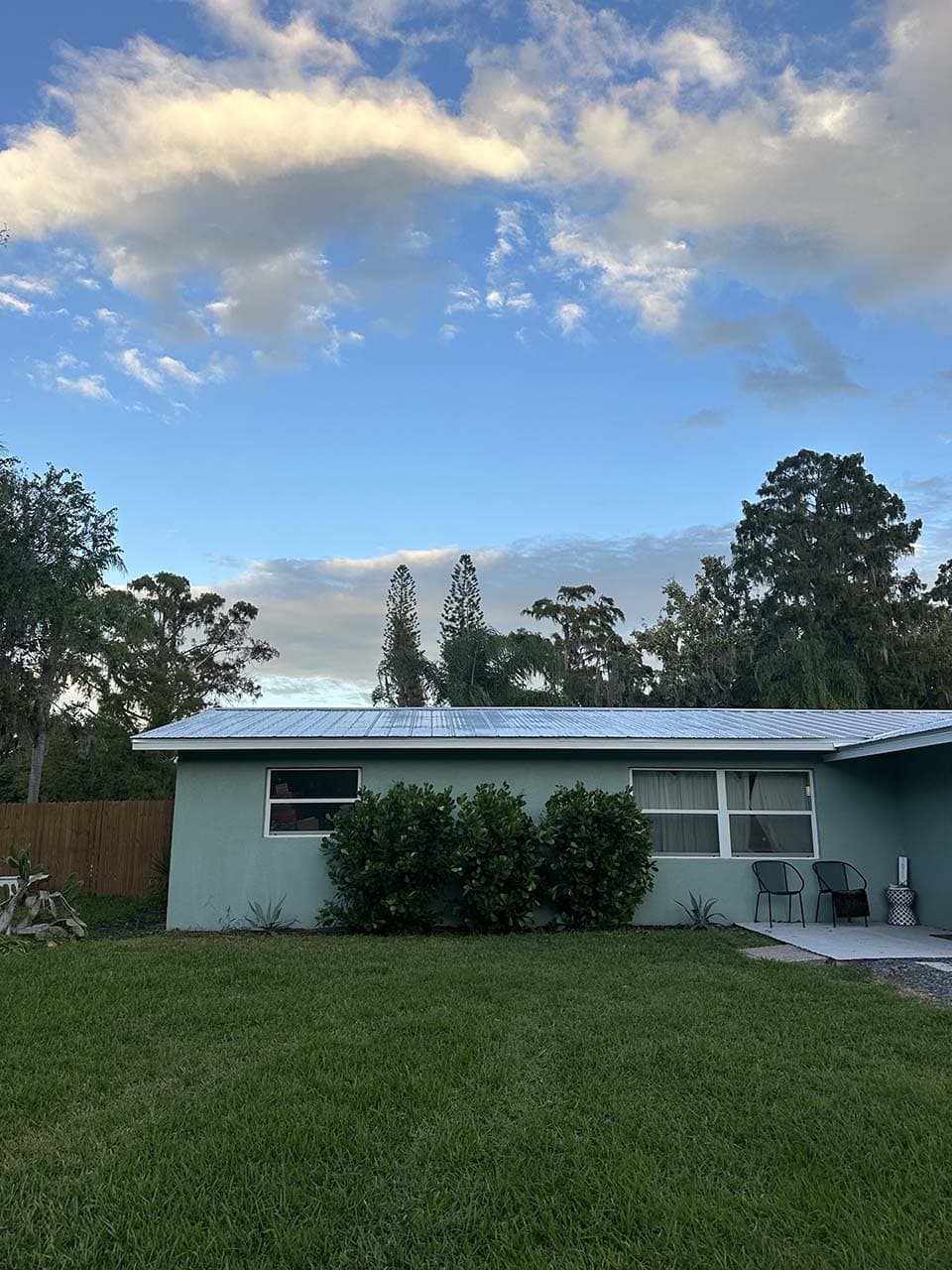 Blue house with white roof, green lawn, and trees under a clear sky with clouds.